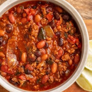 Close-up overhead view of a hearty bowl of spicy chili, filled with ground meat, pinto beans, black beans, tomatoes, and poblano peppers in a rich red sauce, with lime wedges on the side.