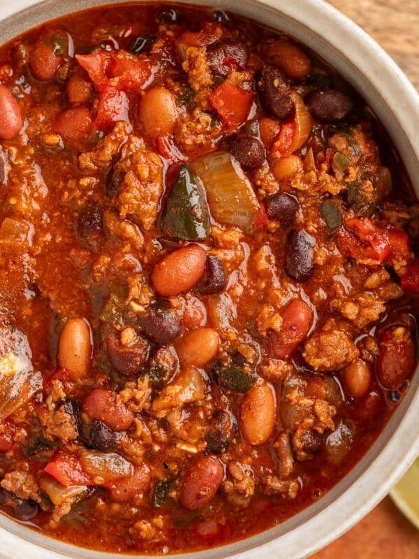 Close-up overhead view of a hearty bowl of spicy chili, filled with ground meat, pinto beans, black beans, tomatoes, and poblano peppers in a rich red sauce, with lime wedges on the side.