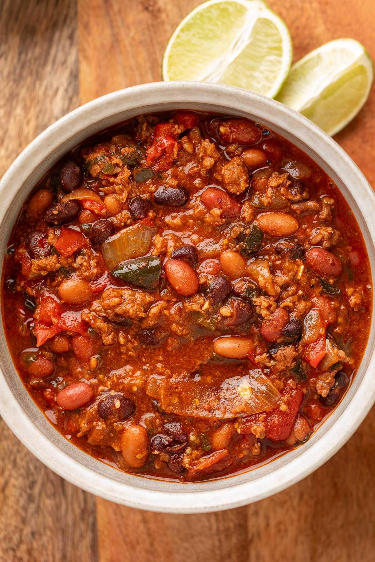 Overhead view of a bowl of spicy chili on a wooden board, filled with ground meat, pinto beans, black beans, tomatoes, and poblano peppers in a thick, red sauce, with lime wedges in the background.