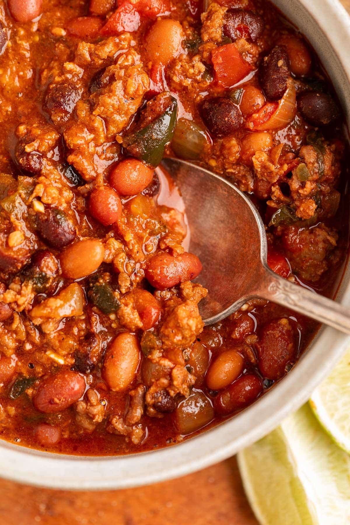 Close-up of a spoonful of spicy chili in a bowl with ground meat, pinto and black beans, tomatoes, and chopped peppers in a thick, red broth.