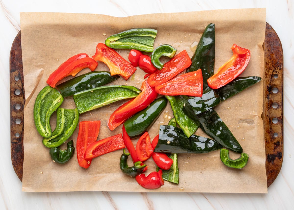 Raw sliced red bell peppers, poblanos, and jalape&ntilde;os arranged on a parchment-lined baking sheet, drizzled with oil and ready for roasting.