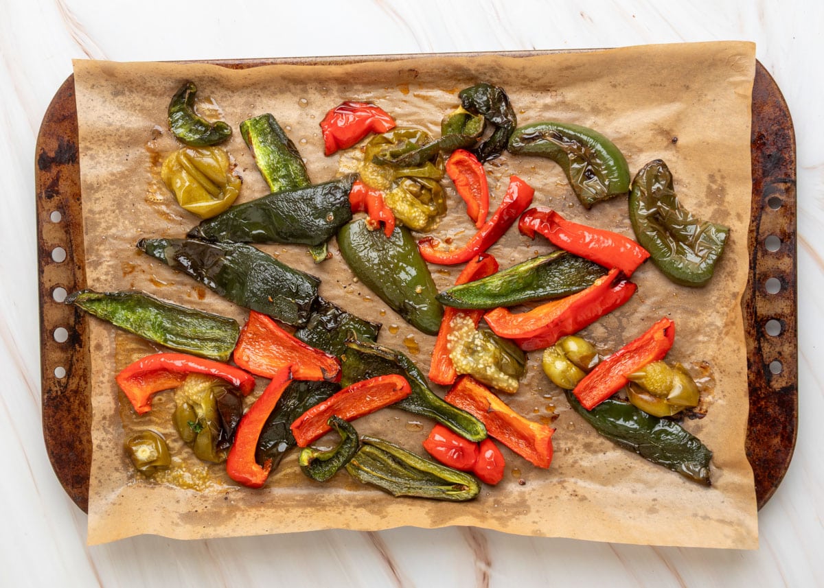 Close-up of roasted peppers and tomatillos on a parchment-lined sheet pan, including blistered red bell peppers, poblanos, jalape&ntilde;os, and softened tomatillos glistening with oil.