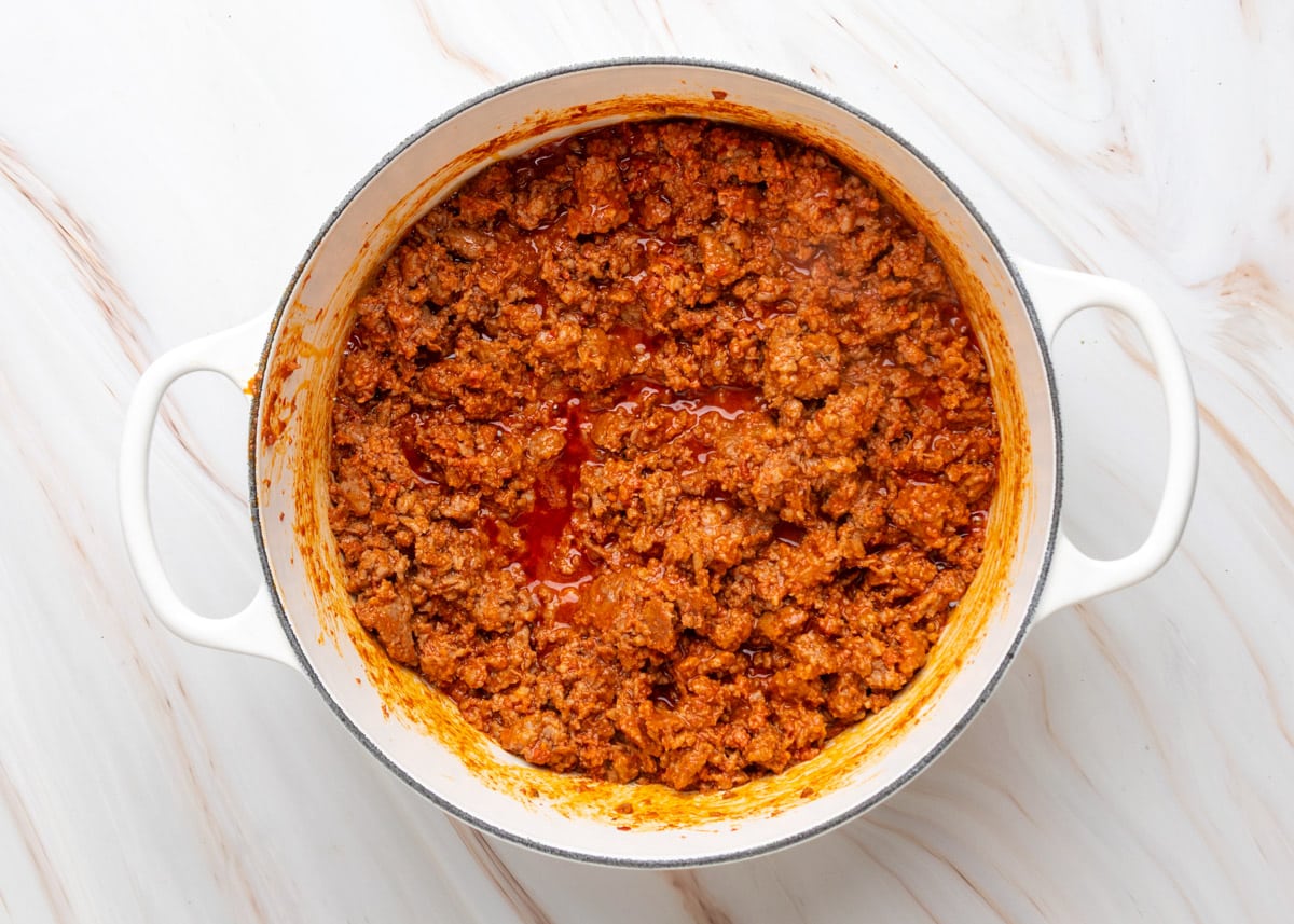 Overhead view of browned ground beef, sausage, and chorizo in a white Dutch oven, coated in a rich red spice blend with rendered fat pooling on the surface.
