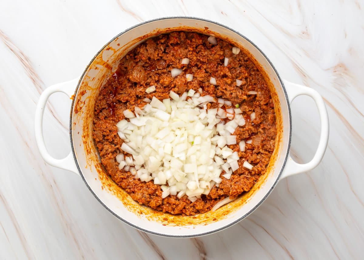 Overhead view of a white Dutch oven filled with browned chili meat, topped with a mound of freshly chopped yellow onions before stirring into the mixture.