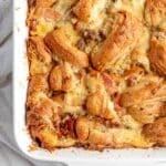 Overhead view of sausage croissant breakfast casserole baked in a white dish, with golden, flaky croissant pieces and melted cheese on top.