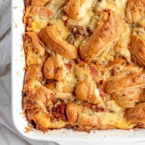 Overhead view of sausage croissant breakfast casserole baked in a white dish, with golden, flaky croissant pieces and melted cheese on top.