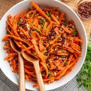 Overhead view of sesame ginger carrot salad with shaved carrots, chili crisp, scallions, chopped peanuts, and sesame seeds in a white serving bowl with wooden salad servers.