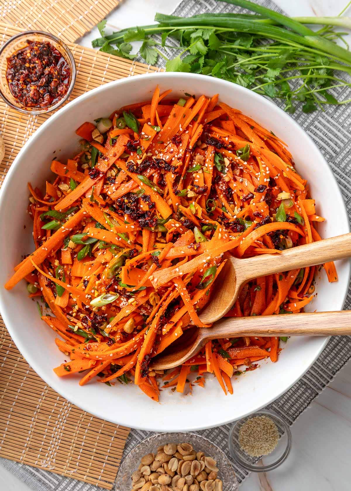 Overhead view of sesame ginger carrot salad made with julienned carrots, scallions, sesame seeds, chili crisp, and chopped peanuts in a white serving bowl with wooden salad servers, surrounded by cilantro, sesame seeds, peanuts, and a small dish of chili crisp.