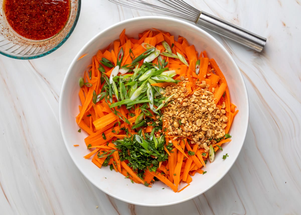 Overhead view of a bowl filled with julienned carrots topped with sliced scallions, chopped cilantro, and crushed peanuts for carrot salad, with a small bowl of chili dressing and a whisk nearby.