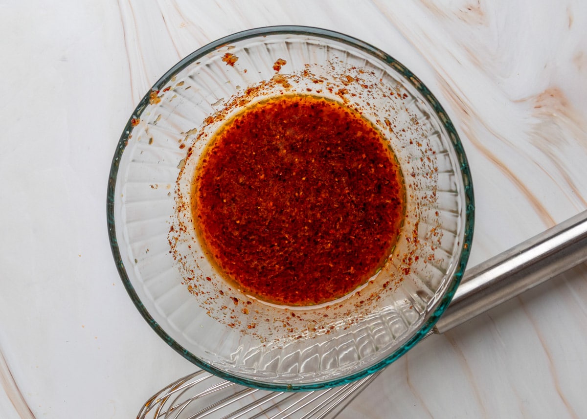 Close-up of a glass bowl containing spicy sesame dressing for carrot salad, showing red chili flakes and oil with a whisk resting alongside.