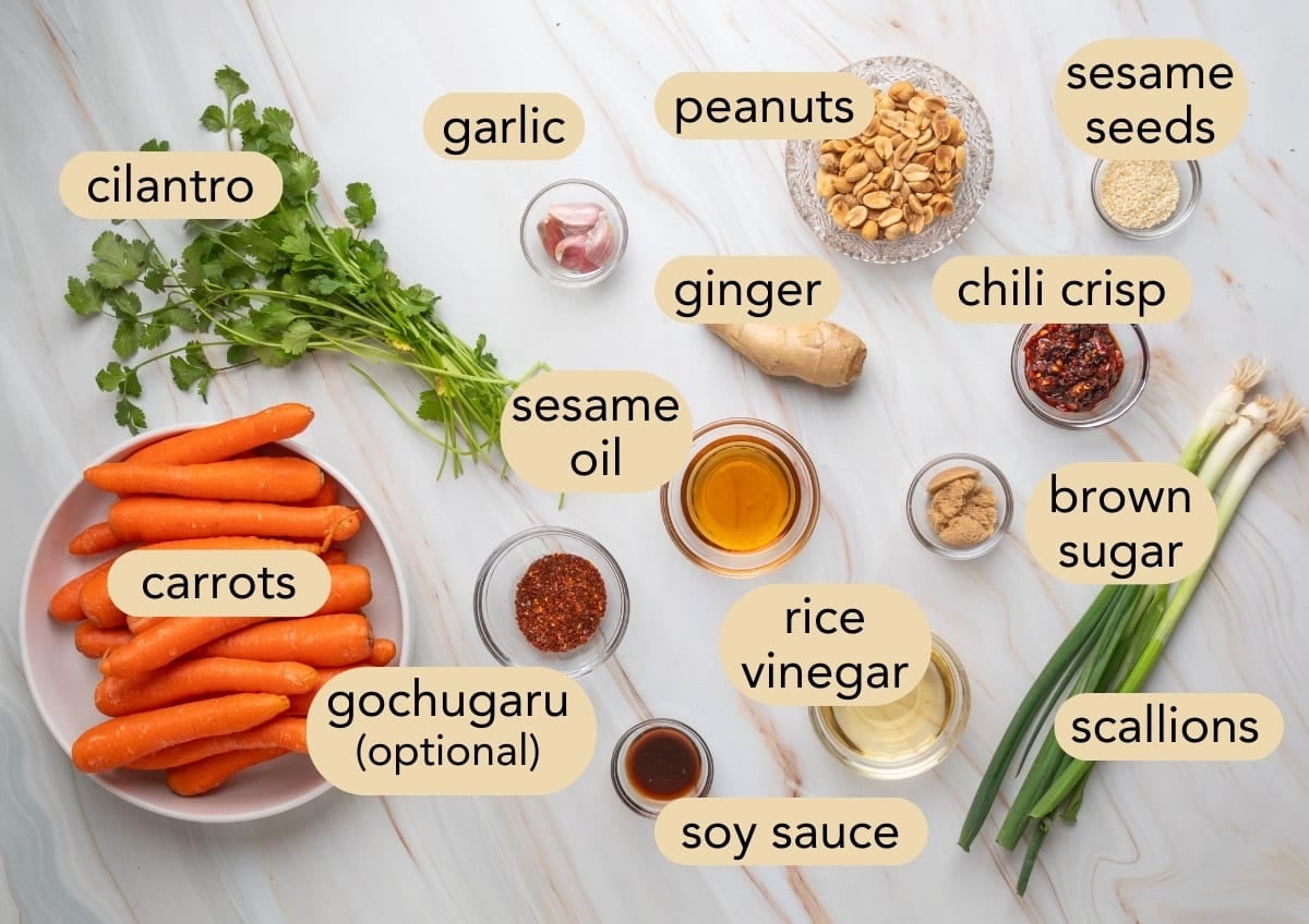 Overhead view of ingredients for sesame ginger carrot salad labeled on the image, including carrots, cilantro, garlic, ginger, scallions, peanuts, sesame seeds, chili crisp, sesame oil, soy sauce, rice vinegar, brown sugar, and optional gochugaru.