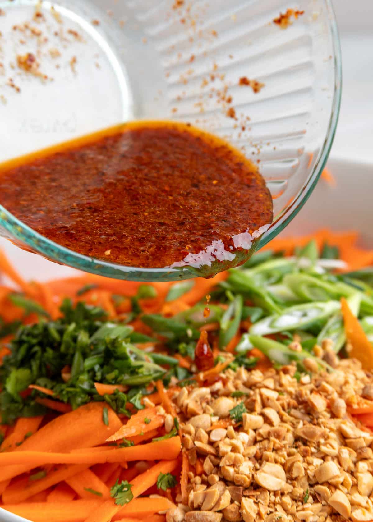 Dressing being poured from a glass bowl over shredded carrots, scallions, chopped peanuts, and cilantro for sesame ginger carrot salad.