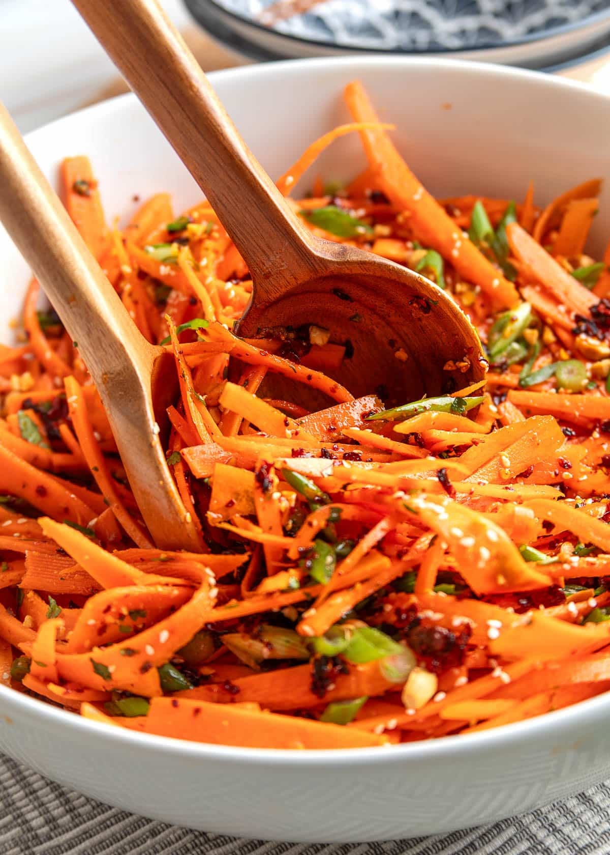 Close-up of sesame ginger carrot salad in a white bowl, with shaved carrots tossed with chili crisp, sesame seeds, scallions, and herbs, being mixed with wooden salad servers.