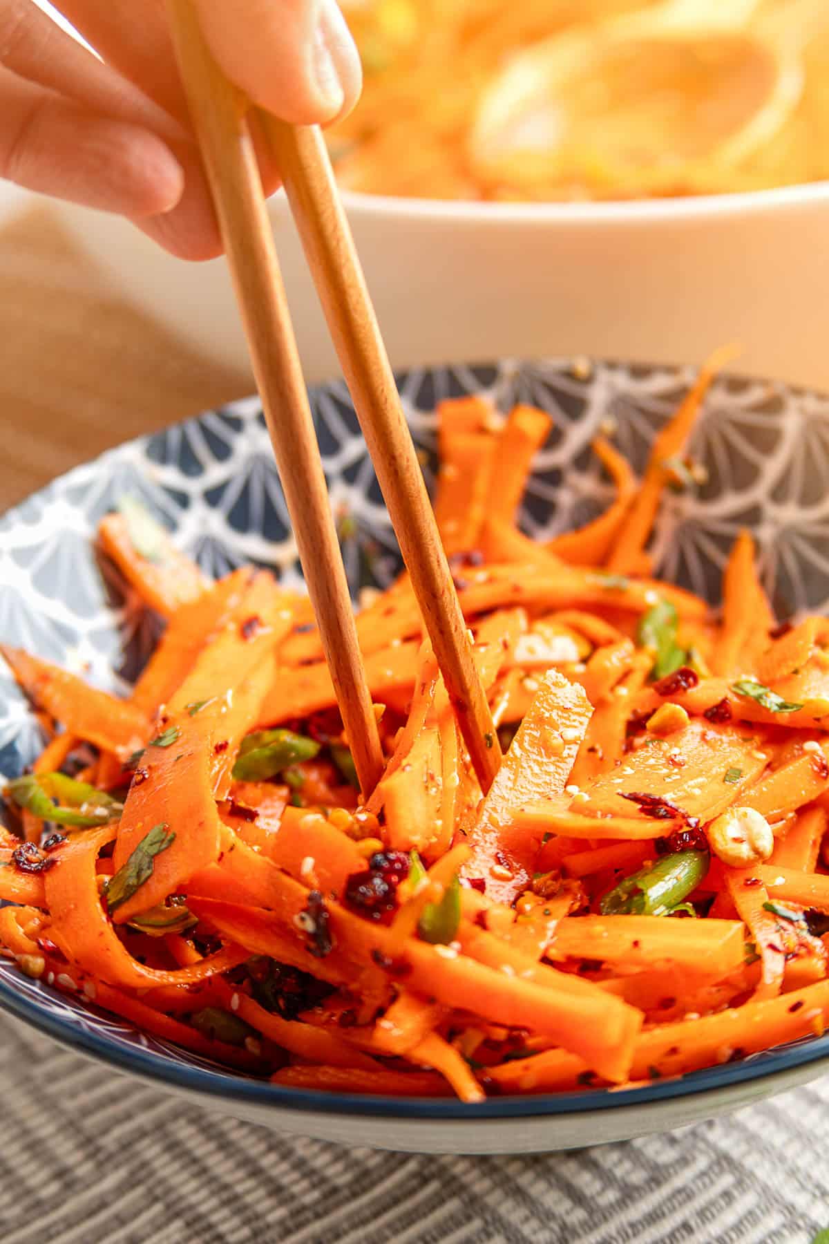 Sesame ginger carrot salad with shaved carrots, chili crisp, scallions, sesame seeds, and peanuts being picked up with chopsticks from a patterned bowl.