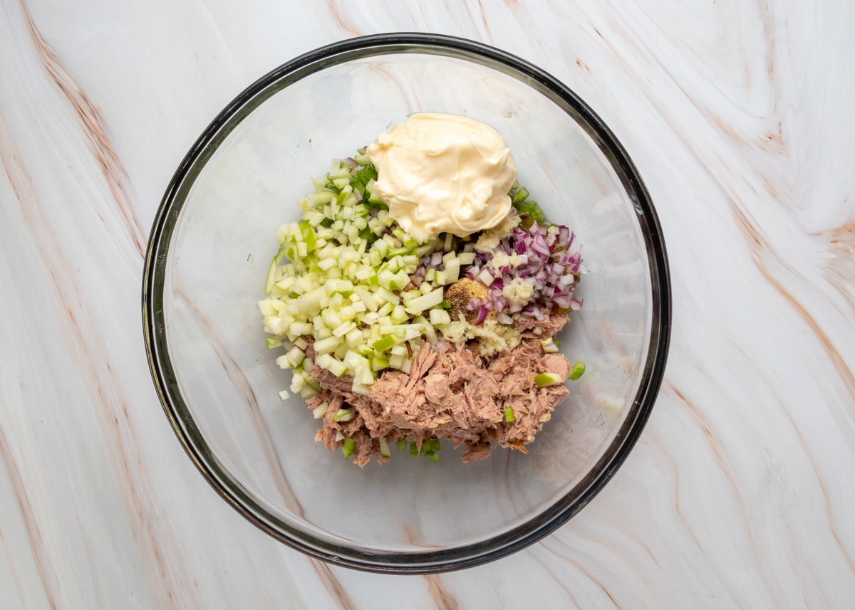 Canned tuna, diced apple, red onion, celery, and mayonnaise in a glass bowl before mixing for apple tuna salad.
