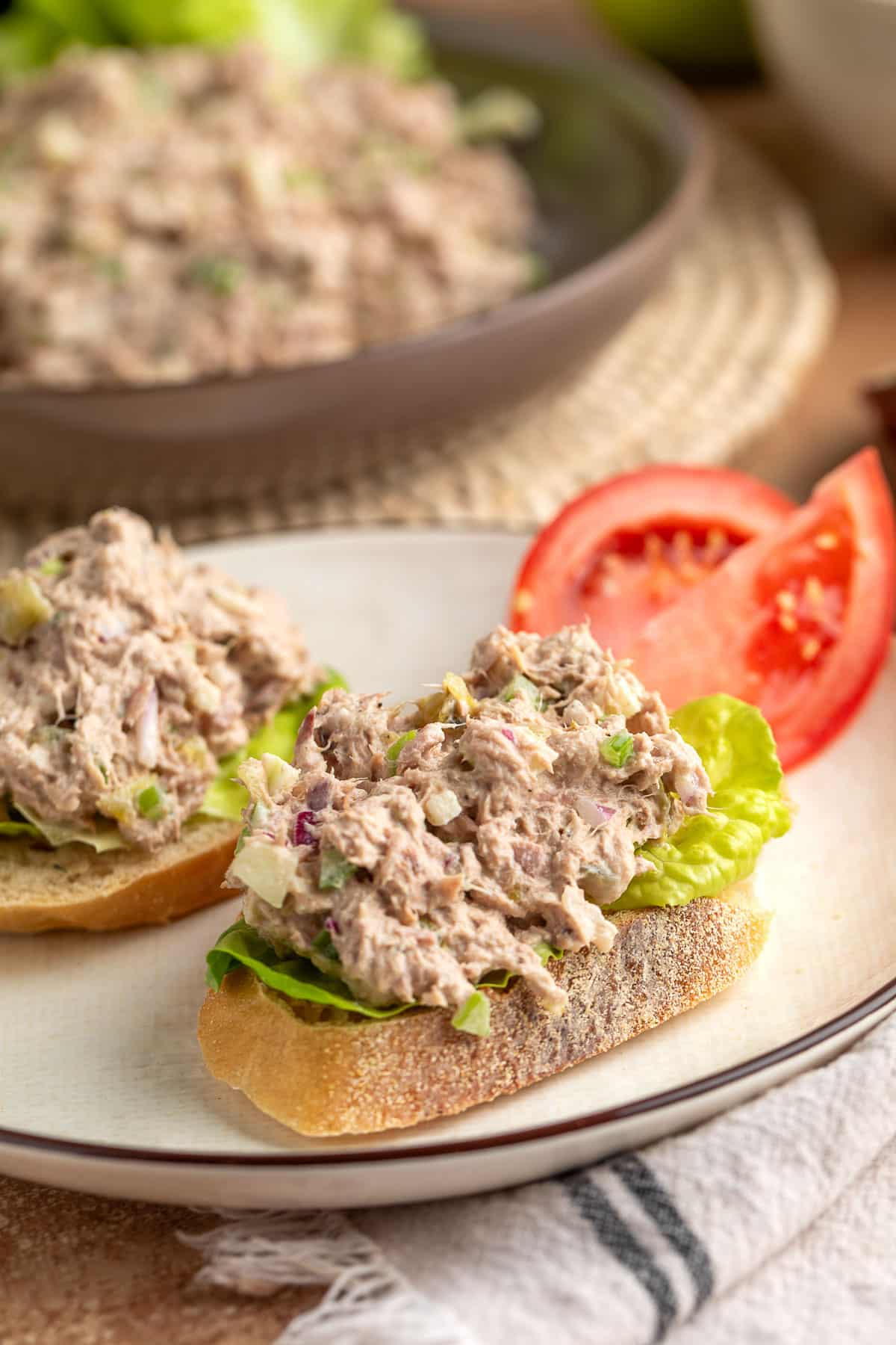 Apple tuna salad served on toasted bread with lettuce, alongside sliced tomato and a bowl of extra salad in the background.