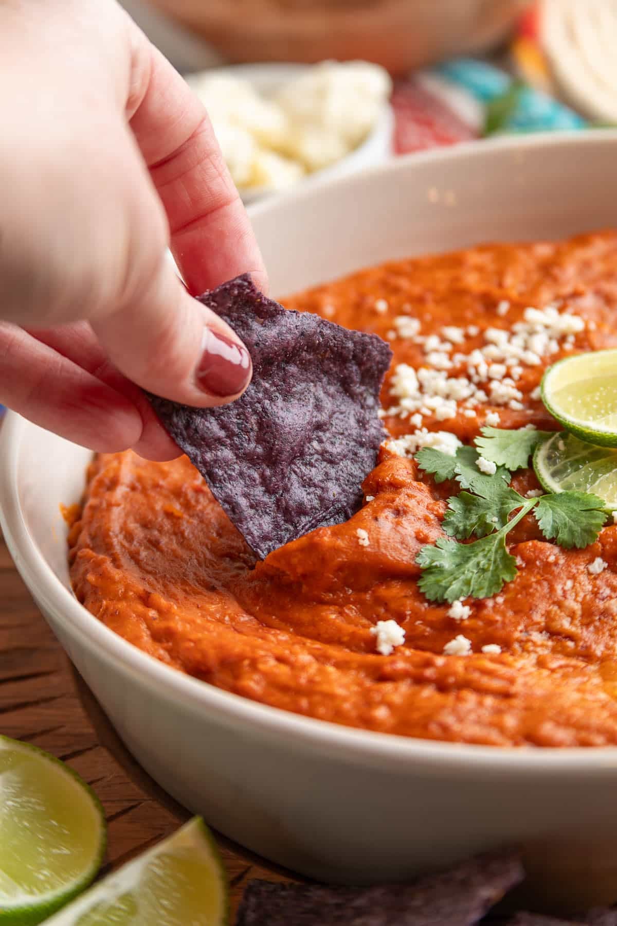 Hand dipping a blue corn tortilla chip into creamy chorizo bean dip topped with crumbled cheese, cilantro, and lime slices.