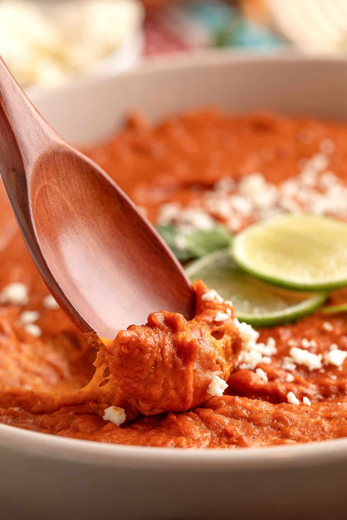 Wooden spoon scooping creamy chorizo bean dip, showing its thick, smooth texture with crumbled cheese and lime slices in the background.