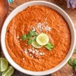 Creamy chorizo bean dip in a white bowl, garnished with crumbled cheese, cilantro, and lime slices, with blue corn tortilla chips and a wooden spoon on the side.