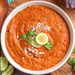 Creamy chorizo bean dip in a white bowl, garnished with crumbled cheese, cilantro, and lime slices, with blue corn tortilla chips and a wooden spoon on the side.