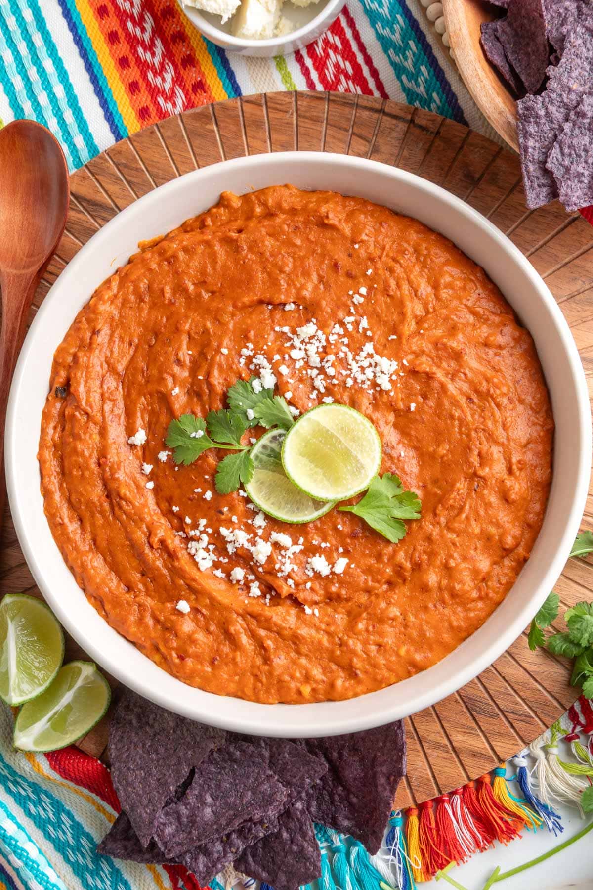 Overhead view of creamy chorizo bean dip in a white bowl, topped with crumbled cheese, lime slices, and cilantro, surrounded by blue corn tortilla chips on a colorful striped cloth.