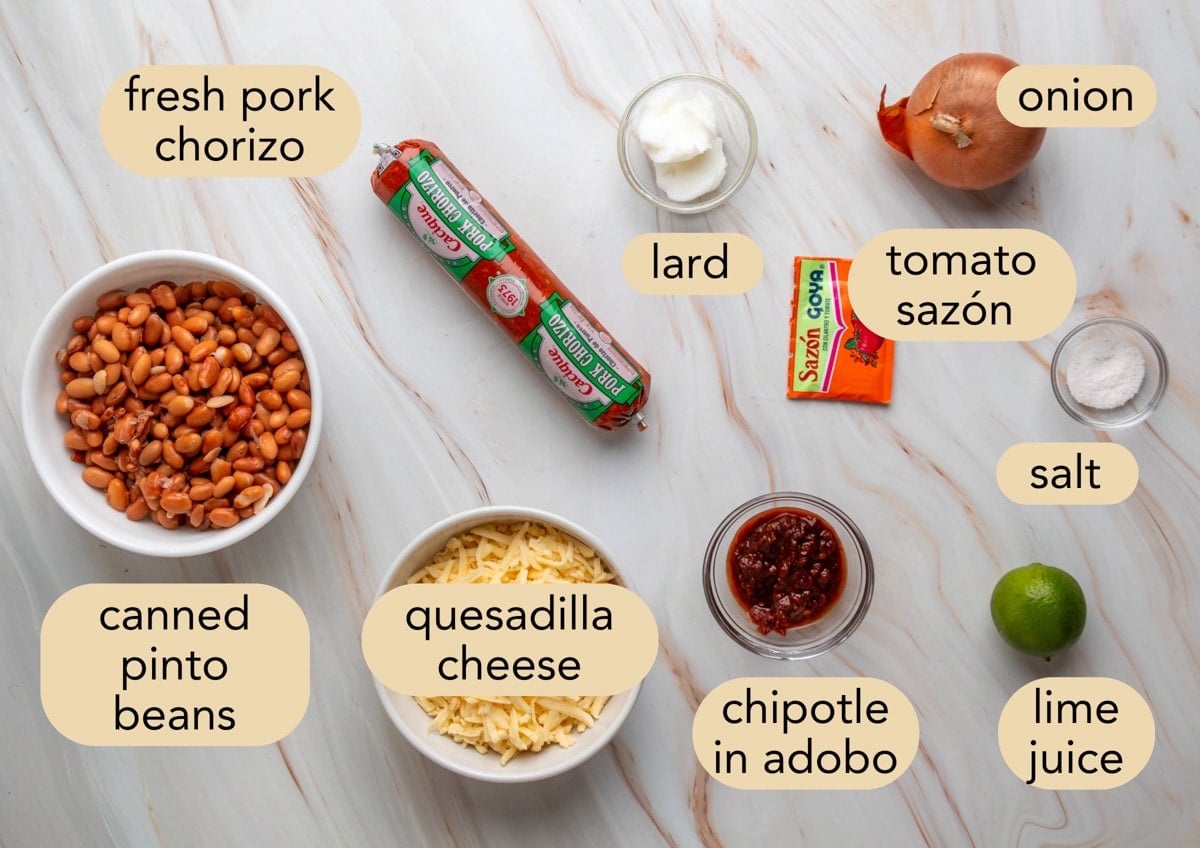 Flat lay of ingredients labeled for chorizo bean dip: fresh pork chorizo, canned pinto beans, shredded quesadilla cheese, chipotle in adobo, lard, tomato saz&oacute;n packet, onion, salt, and lime juice.