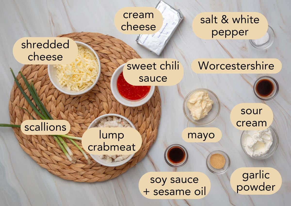 Ingredients for crab rangoon dip labeled on a marble surface, including lump crabmeat, cream cheese, shredded cheese, scallions, sweet chili sauce, mayo, sour cream, Worcestershire sauce, soy sauce with sesame oil, garlic powder, and salt and white pepper.