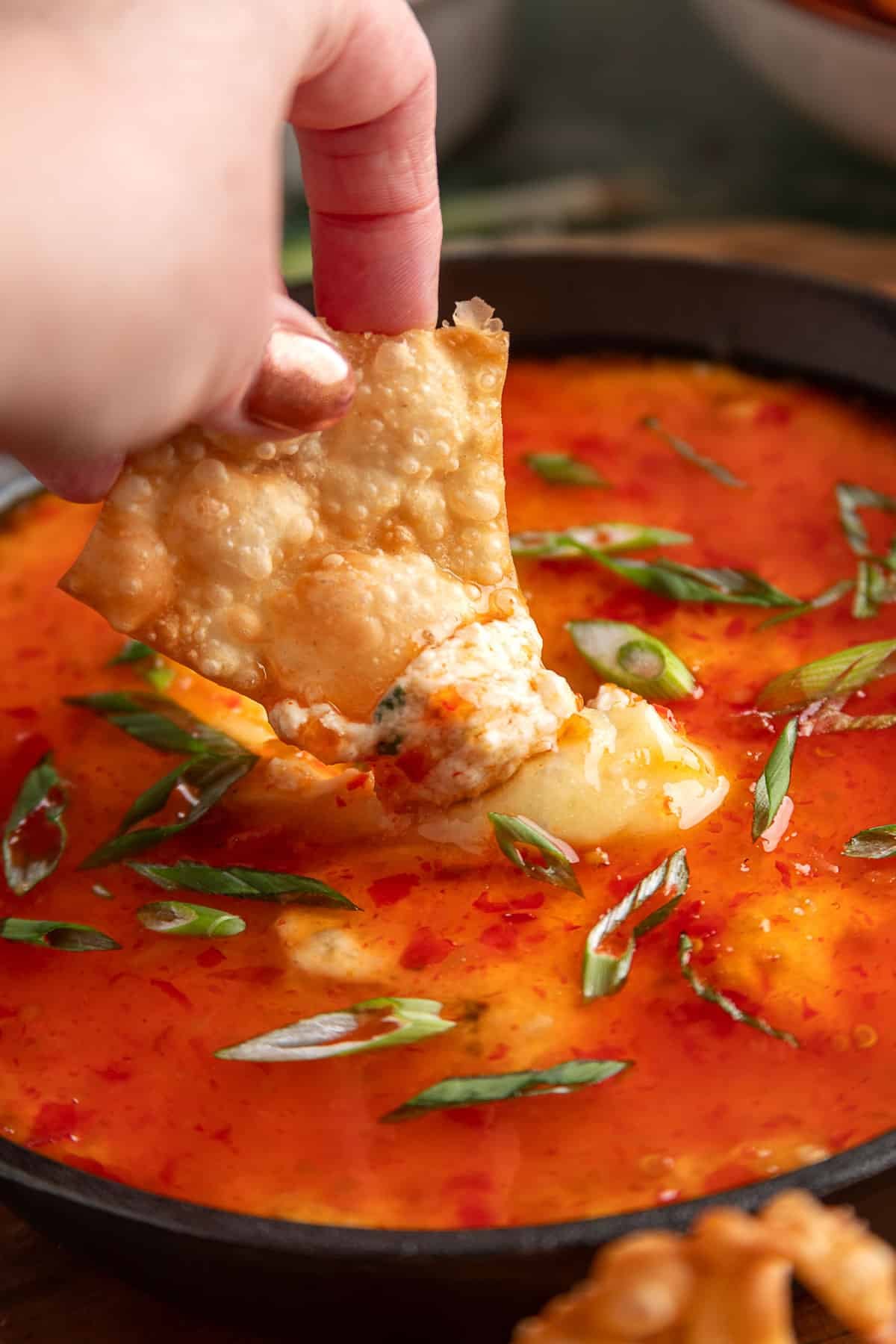 Hand dipping a crispy wonton chip into hot crab rangoon dip topped with sweet chili sauce and sliced green onions.