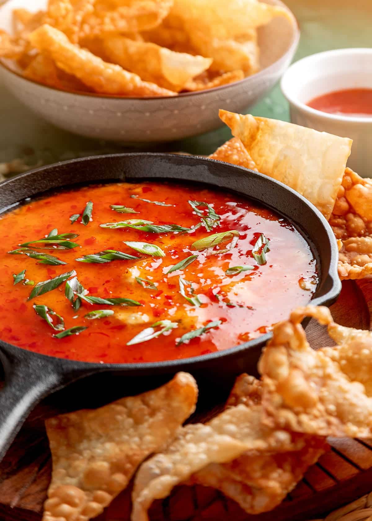 Crab rangoon dip in a cast iron skillet topped with sweet chili sauce and green onions, served with crispy wonton chips on the side.