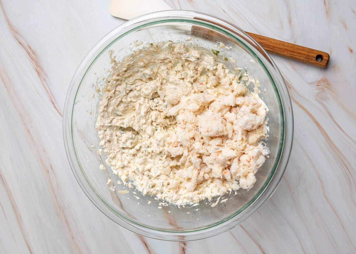 Lump crabmeat being folded into a creamy crab rangoon dip mixture in a glass mixing bowl with a spatula nearby.