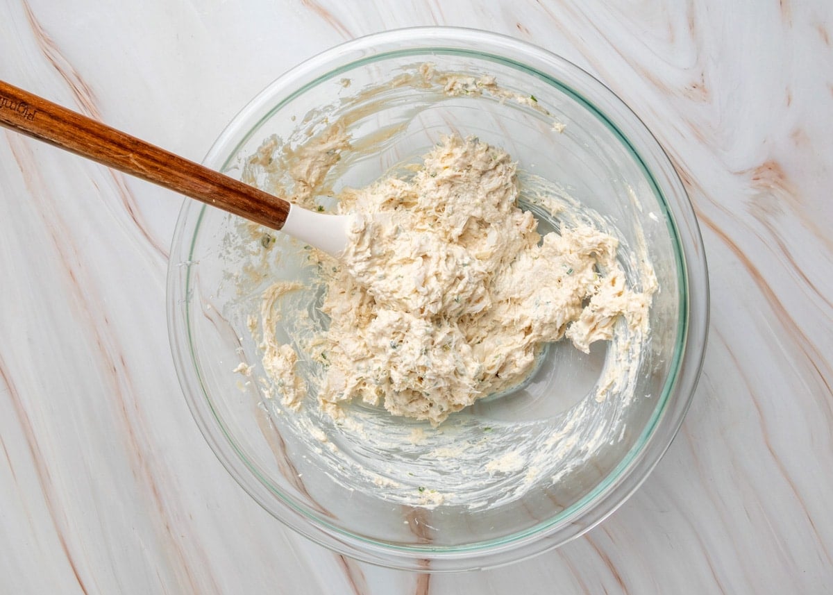 Crab rangoon dip mixture partially combined in a glass bowl with a spatula, showing a thick, creamy texture.