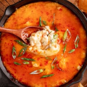 Close-up of crab rangoon dip in a cast iron skillet, topped with sweet chili sauce and sliced green onions, with a wooden spoon scooping up a creamy bite.