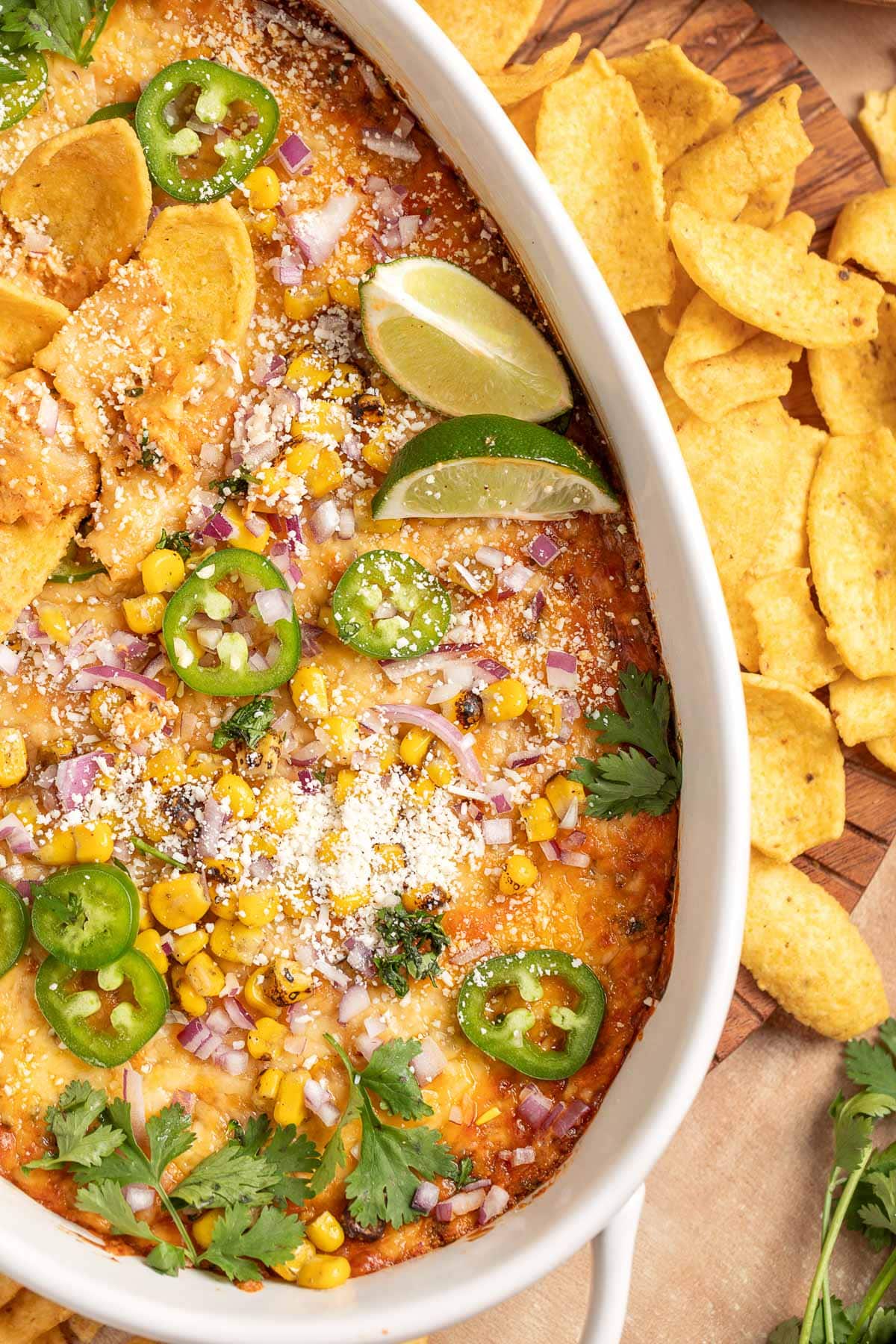 Overhead view of a baked Mexican-style corn dip in a white dish, topped with charred corn, sliced jalape&ntilde;os, diced red onion, cilantro, lime wedges, and crumbled cheese, with tortilla chips on the side.
