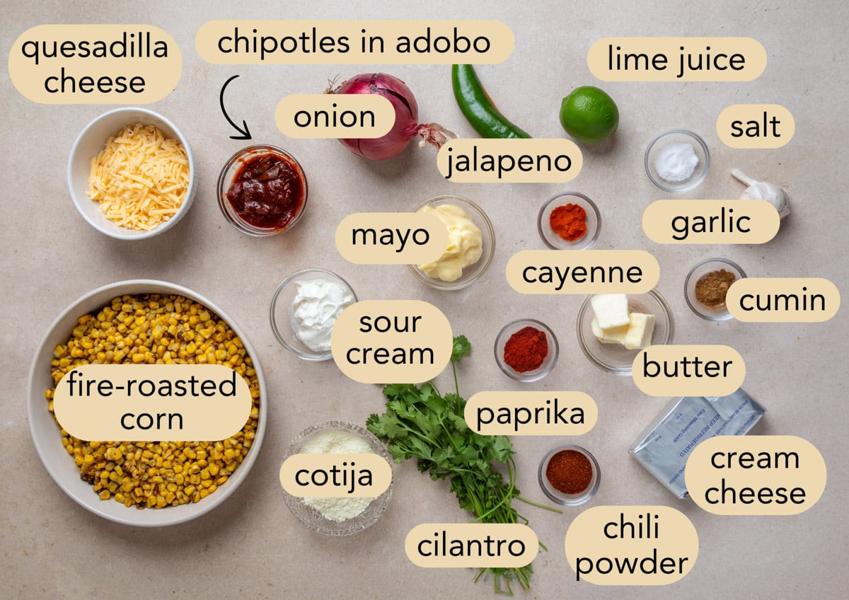 Overhead view of ingredients for Mexican street corn dip arranged on a light surface, including fire-roasted corn, quesadilla cheese, cotija, cream cheese, sour cream, mayo, chipotles in adobo, jalape&ntilde;o, red onion, garlic, lime juice, cilantro, butter, and spices (paprika, chili powder, cumin, cayenne, salt), each labeled.