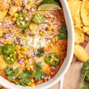 Close-up of baked Mexican street corn dip in a white dish, topped with charred corn, crumbled cotija cheese, diced red onion, sliced jalape&ntilde;os, cilantro, and a lime wedge, with corn chips on the side.