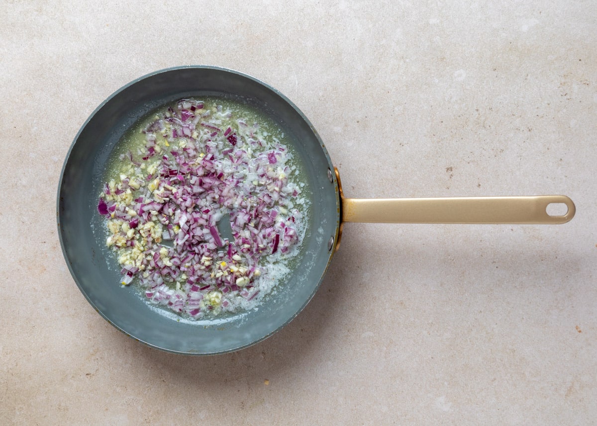 Overhead view of a skillet with finely chopped red onion and garlic saut&eacute;ing in melted butter, just beginning to soften and turn translucent.