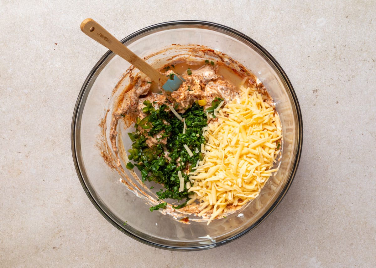 Overhead view of a glass mixing bowl with a creamy base partially mixed with chopped jalape&ntilde;os, cilantro, and shredded cheese, with a spatula resting in the bowl.