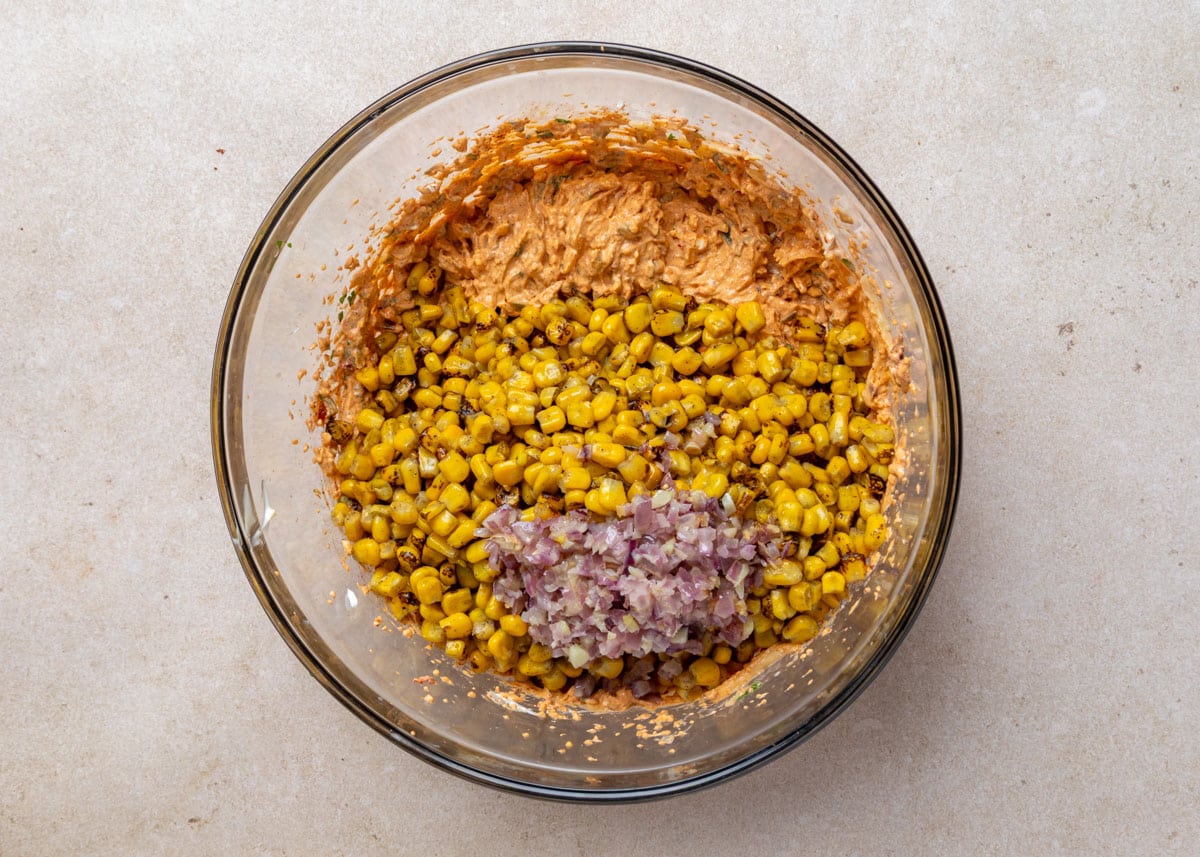 Overhead view of a glass bowl with a creamy, seasoned base mixed with charred corn and sauteed finely diced red onion, ready to be stirred together.