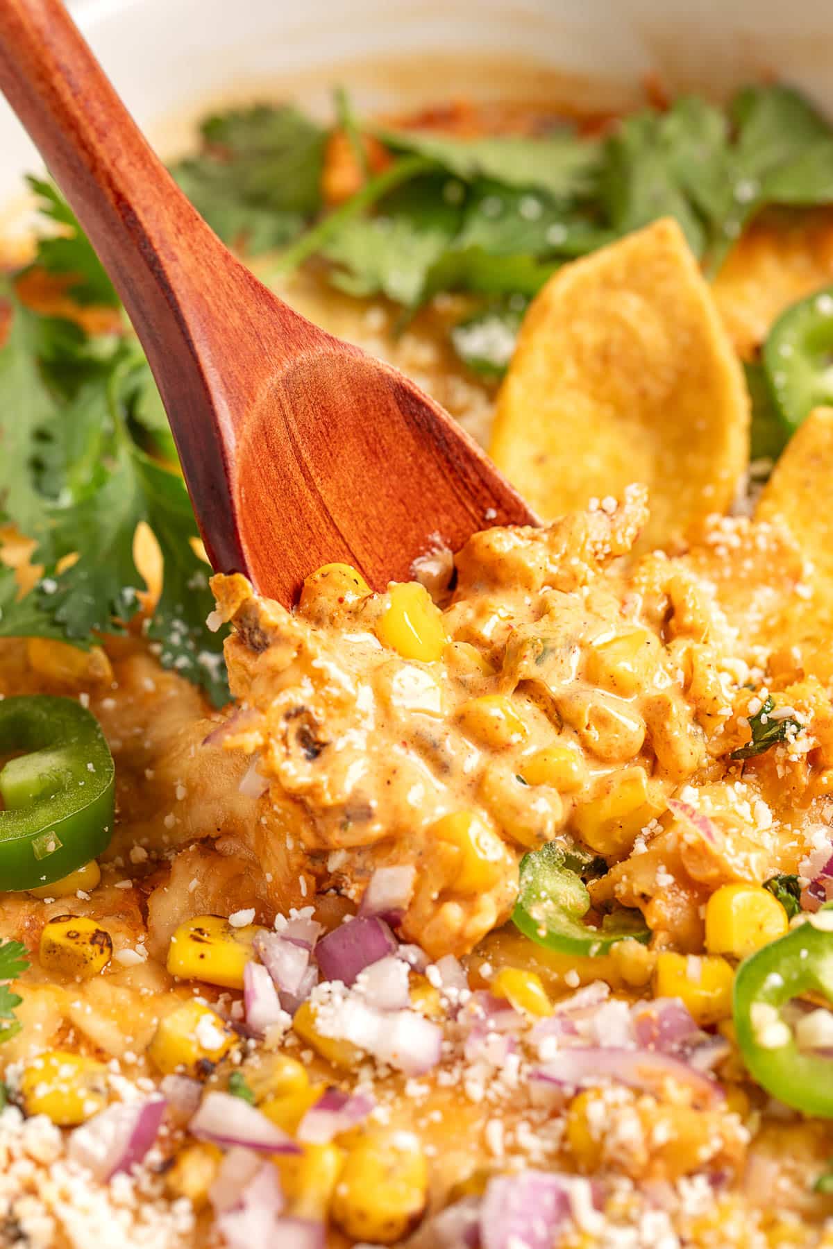 Close-up of a spoon scooping creamy Mexican street corn dip, with charred corn, diced red onion, jalape&ntilde;o slices, cilantro, and crumbled cheese visible, alongside tortilla chips.