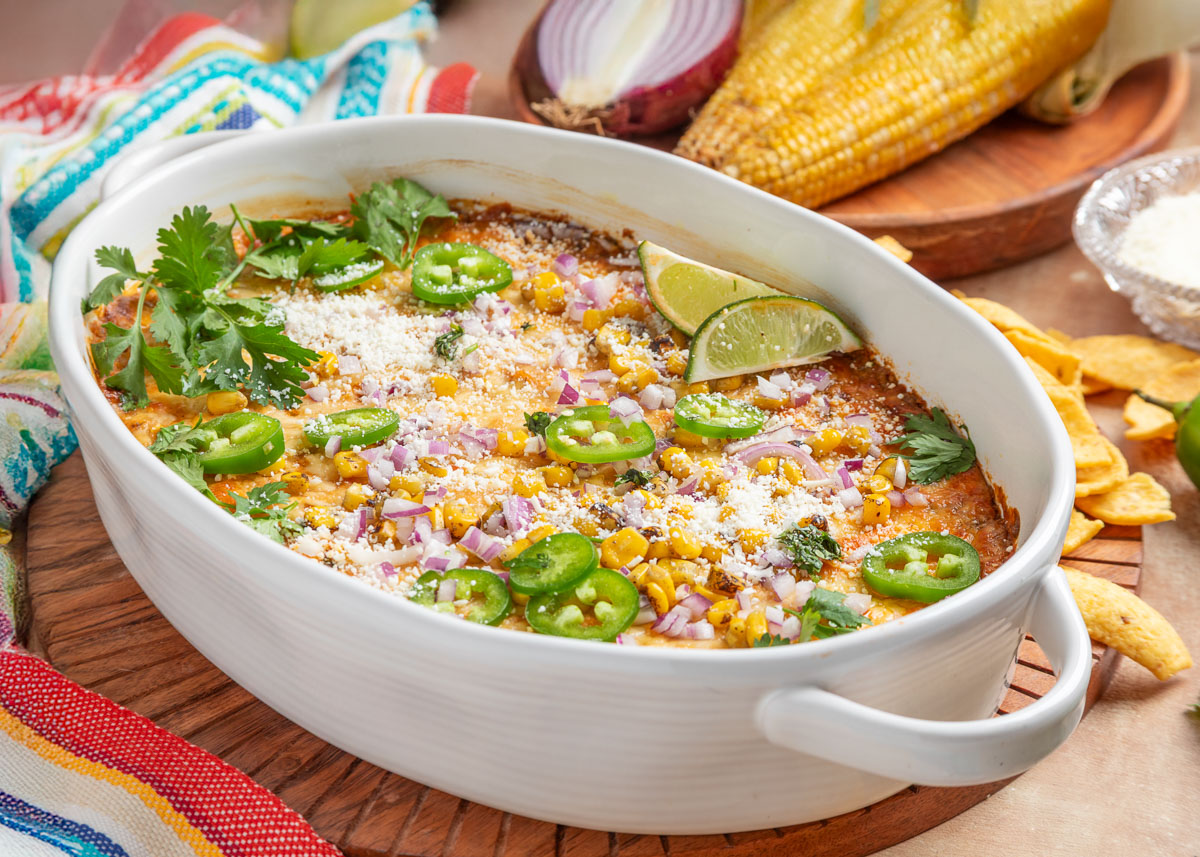 Baked Mexican street corn dip in a white casserole dish, topped with jalape&ntilde;o slices, corn, diced red onion, cilantro, lime wedges, and crumbled cheese, with tortilla chips and fresh ingredients in the background.