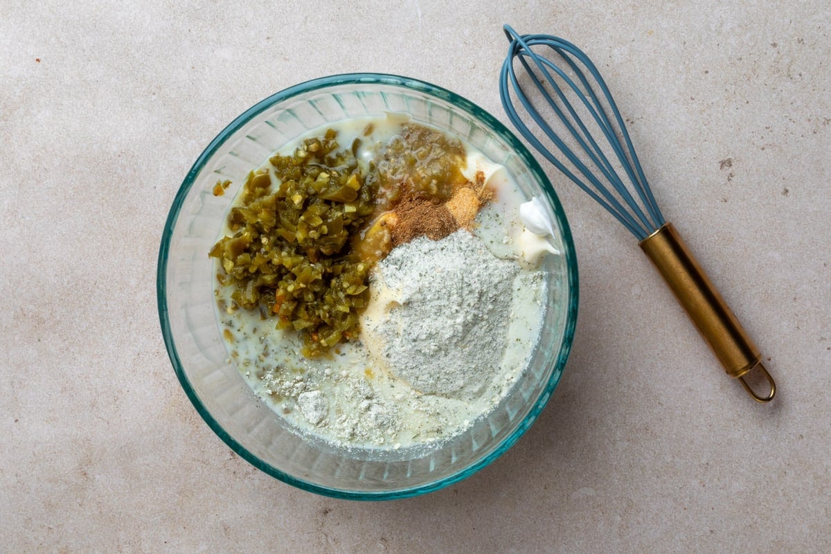 Overhead photo of ingredients for cilantro jalape&ntilde;o dressing combined in a glass mixing bowl, including mayonnaise, buttermilk, ranch seasoning, salsa verde, spices, and chopped pickled jalape&ntilde;os, with a whisk alongside.