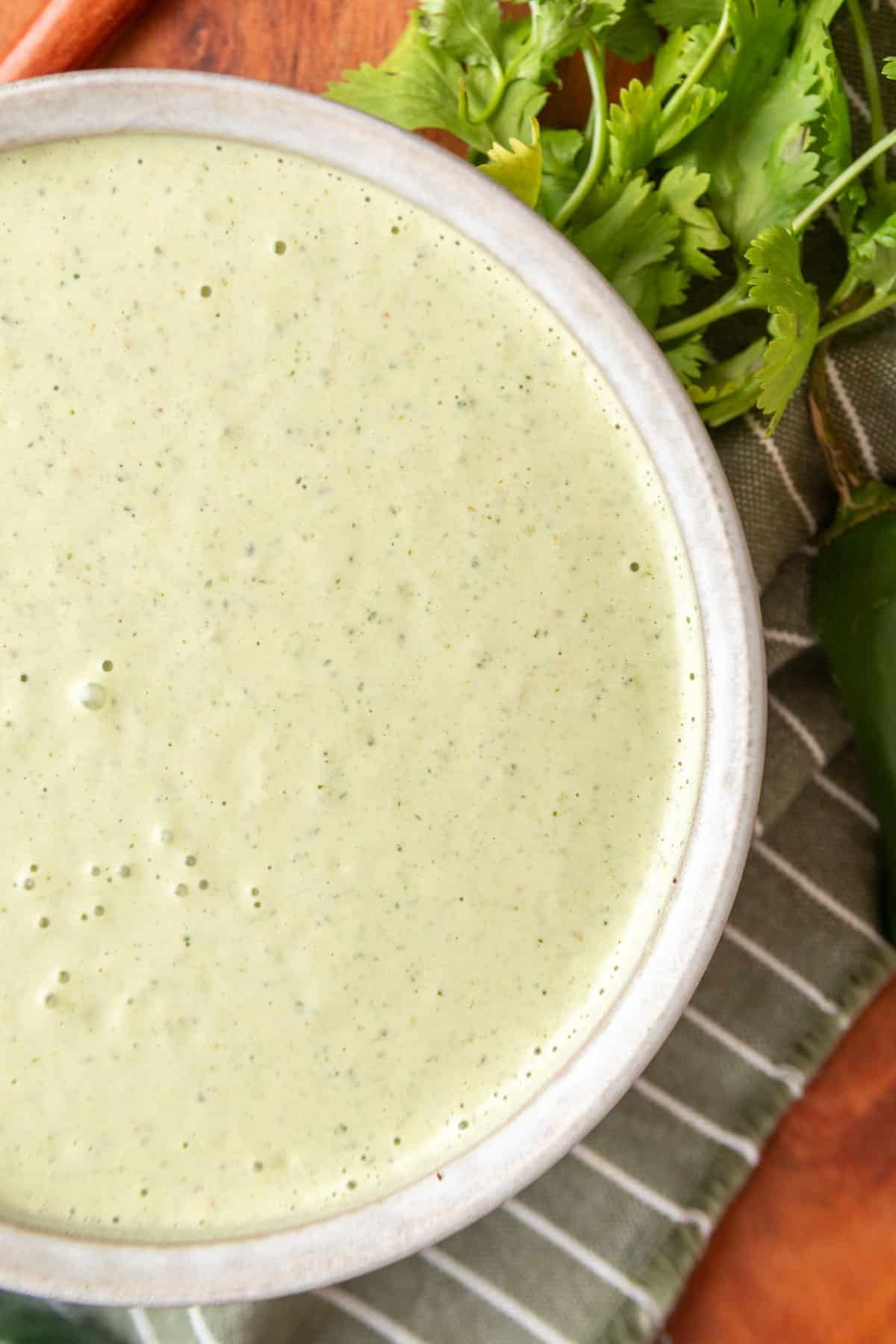 Close-up overhead photo of creamy cilantro jalapeno dressing in a ceramic bowl, with fresh cilantro and jalapenos on the side.