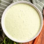 Overhead view of creamy cilantro jalapeño dressing in a shallow stoneware bowl, with a smooth pale green texture and flecks of herbs, styled with fresh cilantro, a striped green towel, and a wooden serving board.