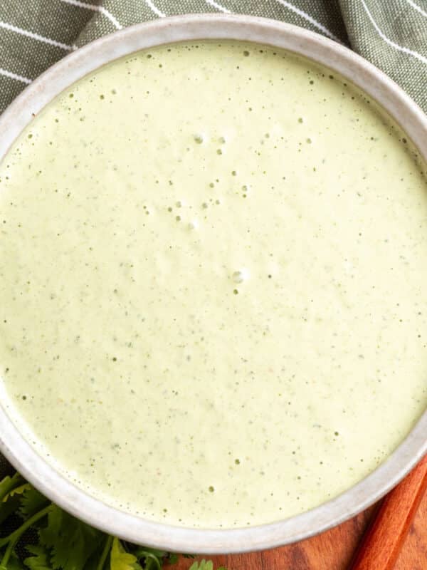 Overhead view of creamy cilantro jalape&ntilde;o dressing in a shallow stoneware bowl, with a smooth pale green texture and flecks of herbs, styled with fresh cilantro, a striped green towel, and a wooden serving board.