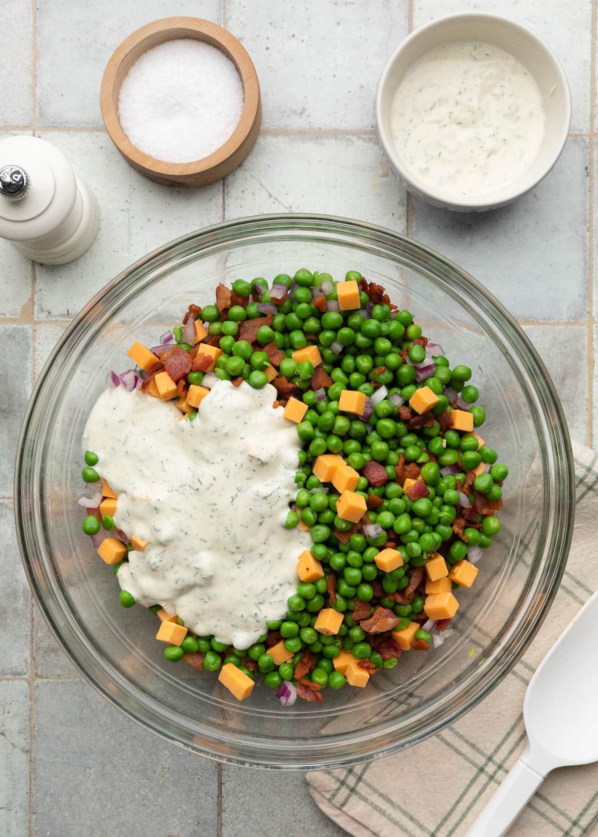 Top-down process photo of creamy dill dressing poured over a bowl of green peas, crispy bacon, cheddar cheese cubes, and diced red onion before mixing.