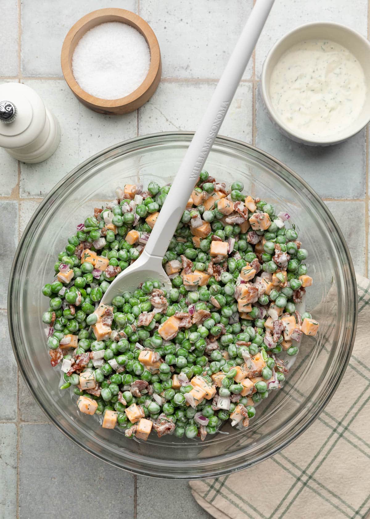 Top-down photo of creamy pea salad tossed in a glass mixing bowl with bright green peas, crispy bacon pieces, cheddar cheese cubes, diced red onion, and dill dressing, with a white rubber spatula resting in the bowl.