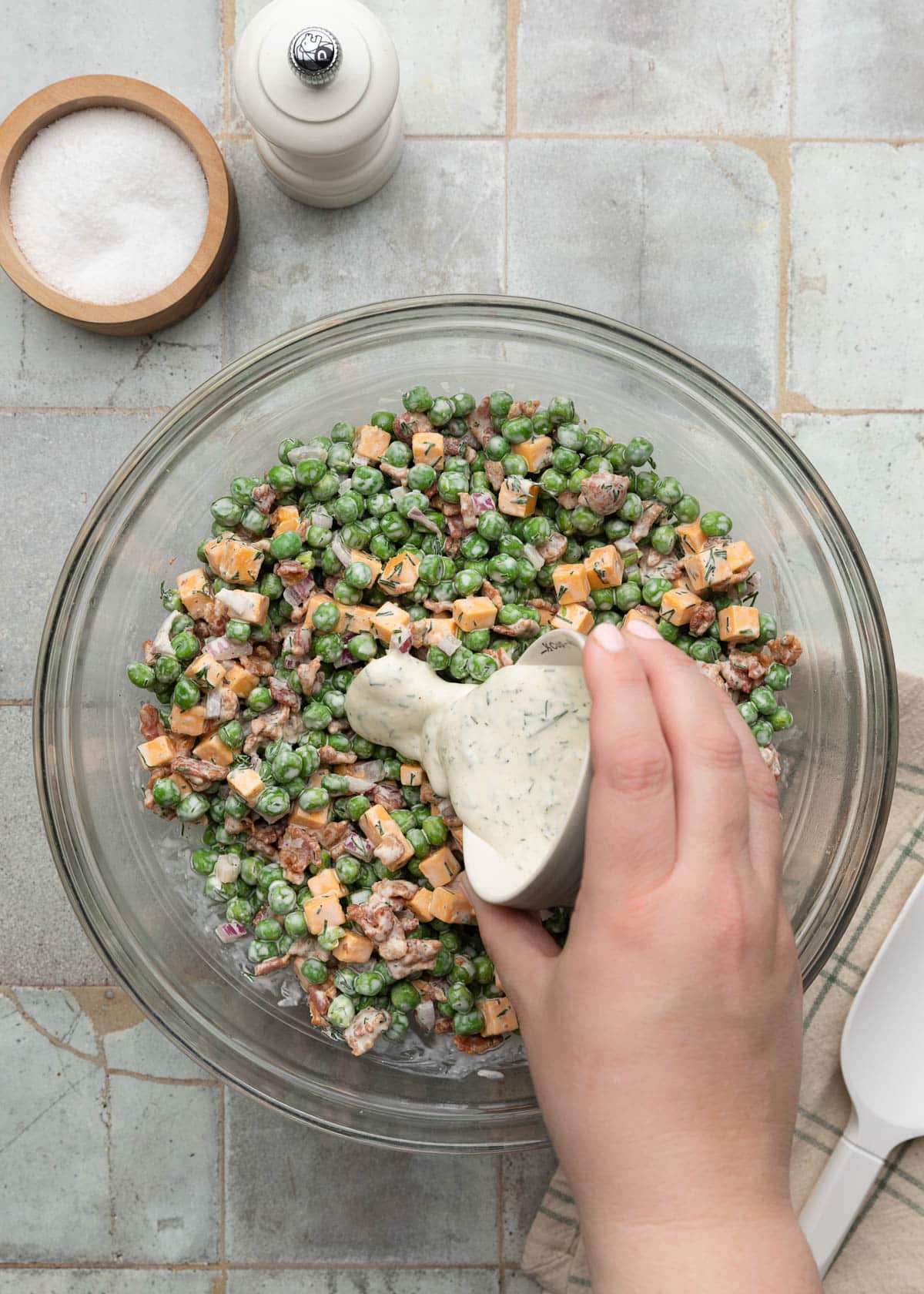 Top-down action photo showing creamy dill dressing being poured from a small bowl over a pea salad made with green peas, cheddar cheese cubes, crispy bacon, and diced red onion in a large glass mixing bowl.