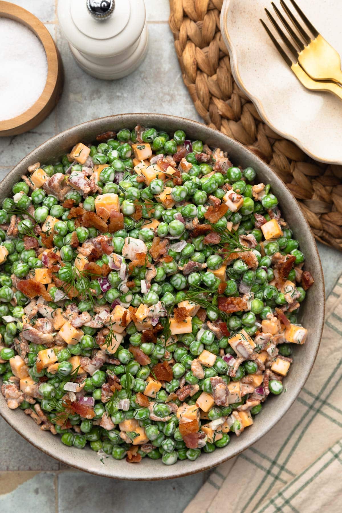 Top-down photo of creamy pea salad in a ceramic serving bowl, made with green peas, cheddar cheese cubes, crispy bacon, diced red onion, and fresh dill tossed in a creamy dressing. A white pepper mill, salt cellar, woven placemat, and gold fork are arranged around the bowl on a tiled surface.