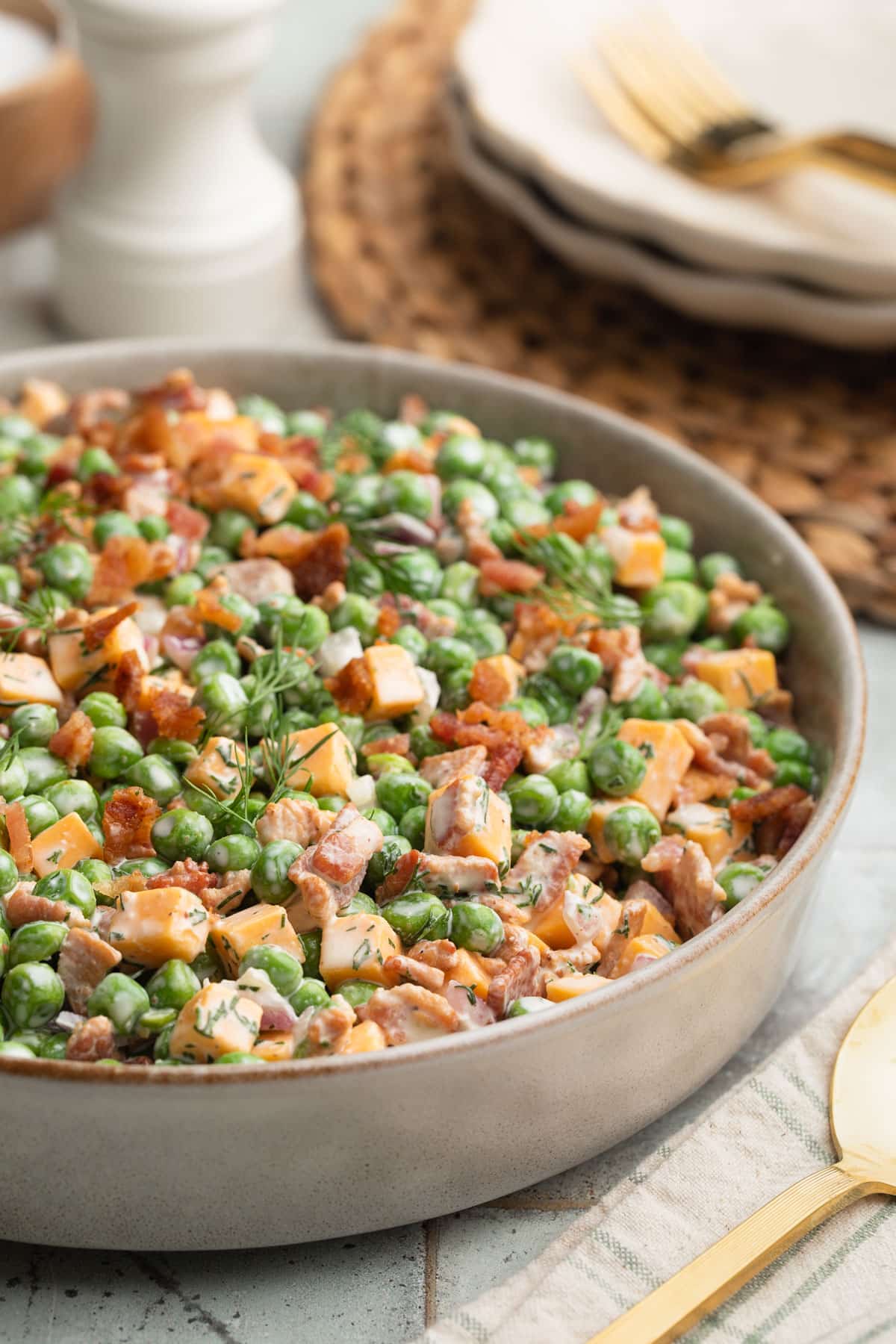 Close-up photo of creamy pea salad served in a shallow ceramic bowl, made with green peas, cheddar cheese cubes, crispy bacon, chopped pecans, diced red onion, and fresh dill tossed in a creamy dressing. Gold flatware and stacked plates sit blurred in the background.