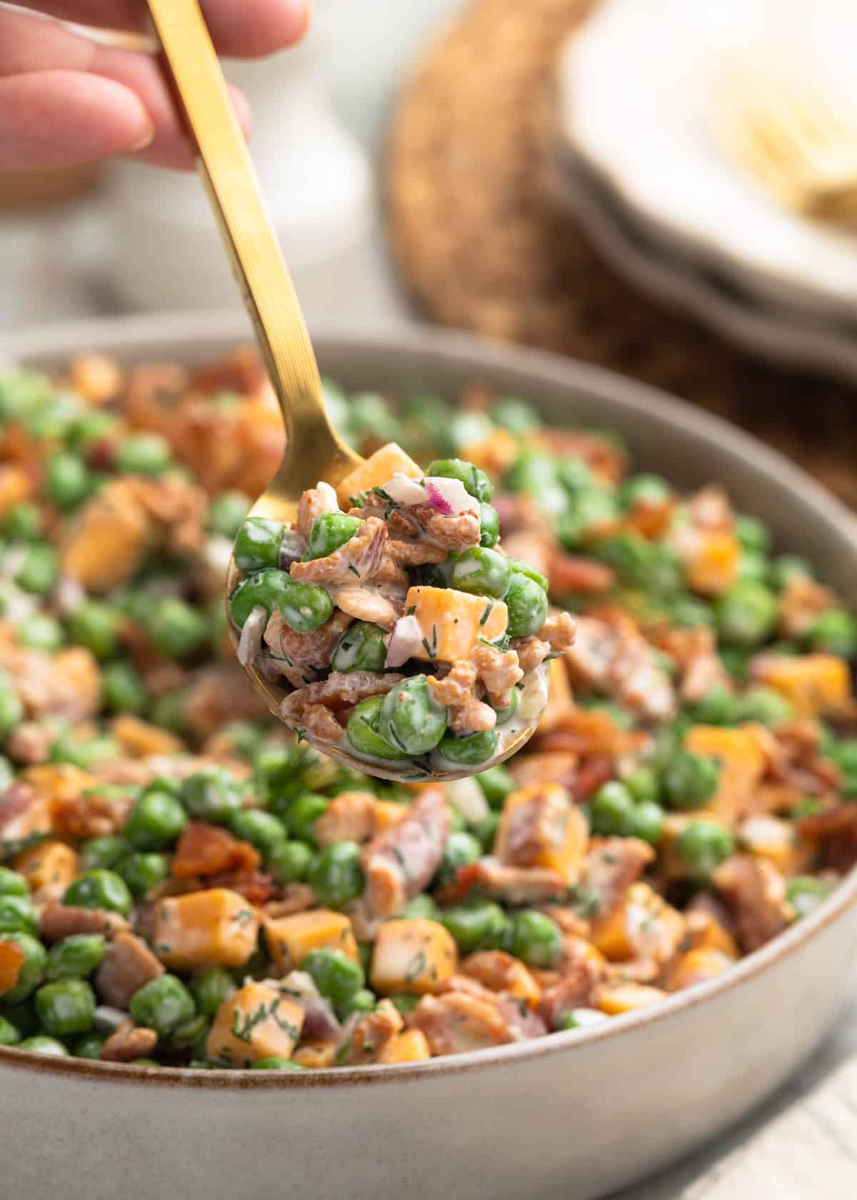 Close-up photo of a spoonful of creamy pea salad with green peas, cheddar cheese cubes, crispy bacon, diced red onion, and dill dressing held above a serving bowl filled with the salad.